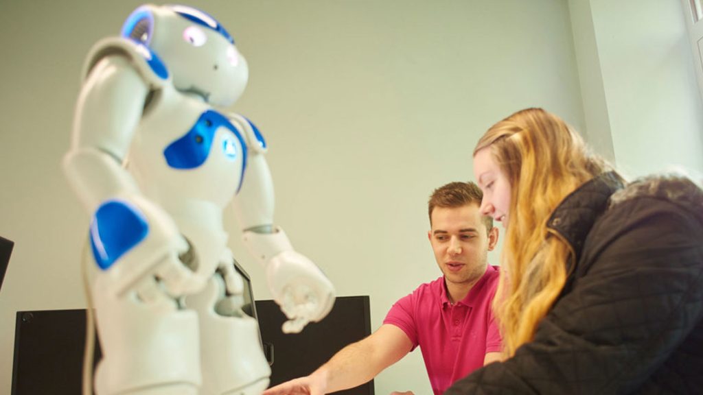 Two students working on a computer with a robot in the foreground