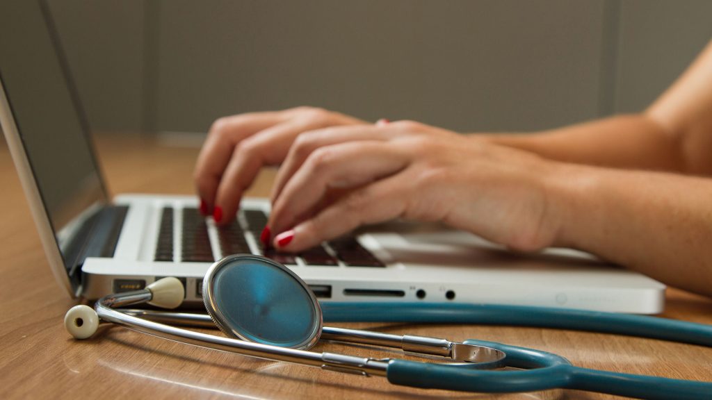 a persons hands on their laptop, typing. They have a stethoscope next to their laptop on their desk.