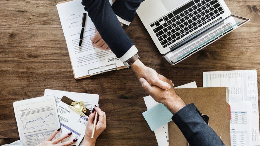 Birds eye view of two people shaking hands over a desk, laptop and paperwork