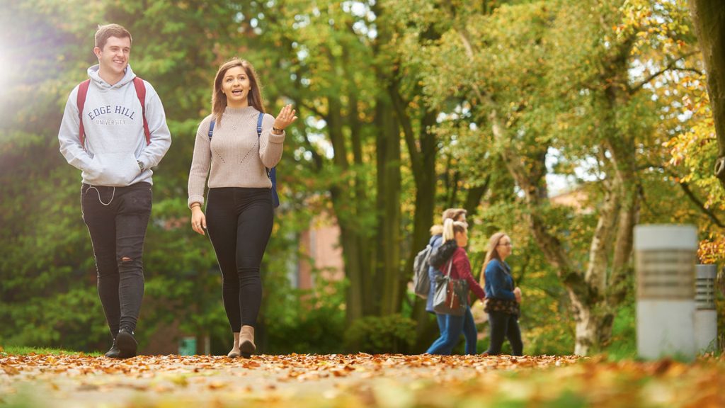 Two students walking through campus in Autumn. There are leaves on the pavement and the trees are turning brown in the background.