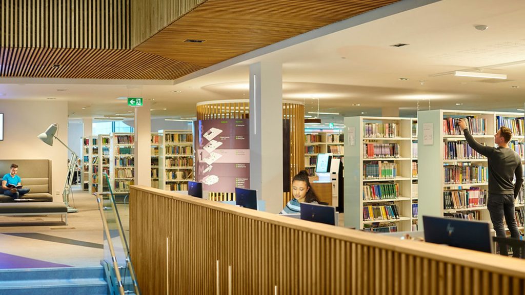 A student is working at a computer in the Catalyst. There is a student in the background picking books from the bookshelves in the background. Another student is reading a book on the couch in the background.