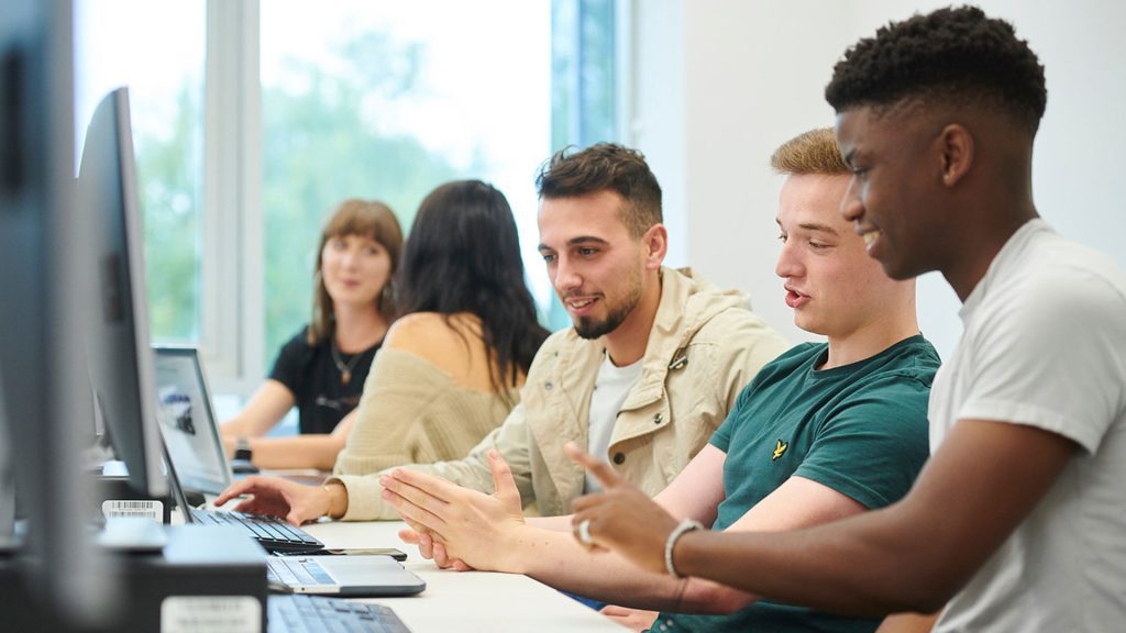 Three students sitting at a bank of computers whilst looking at the same laptop together. There are two students in the background sitting together and chatting, whilst also on laptops.