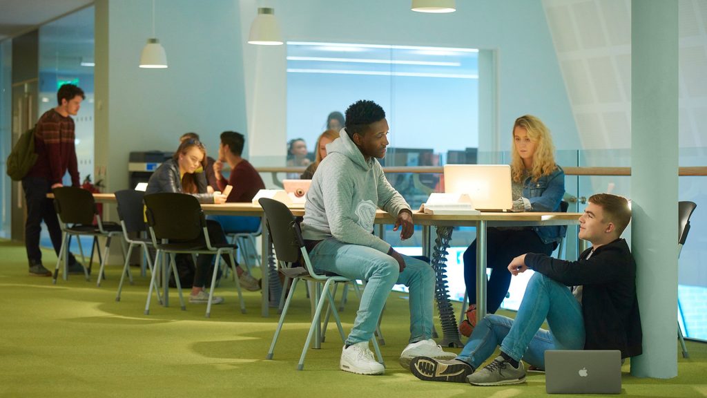 8 students studying across 4 different tables. Two students in conversation at the focal point of the image with a laptop.