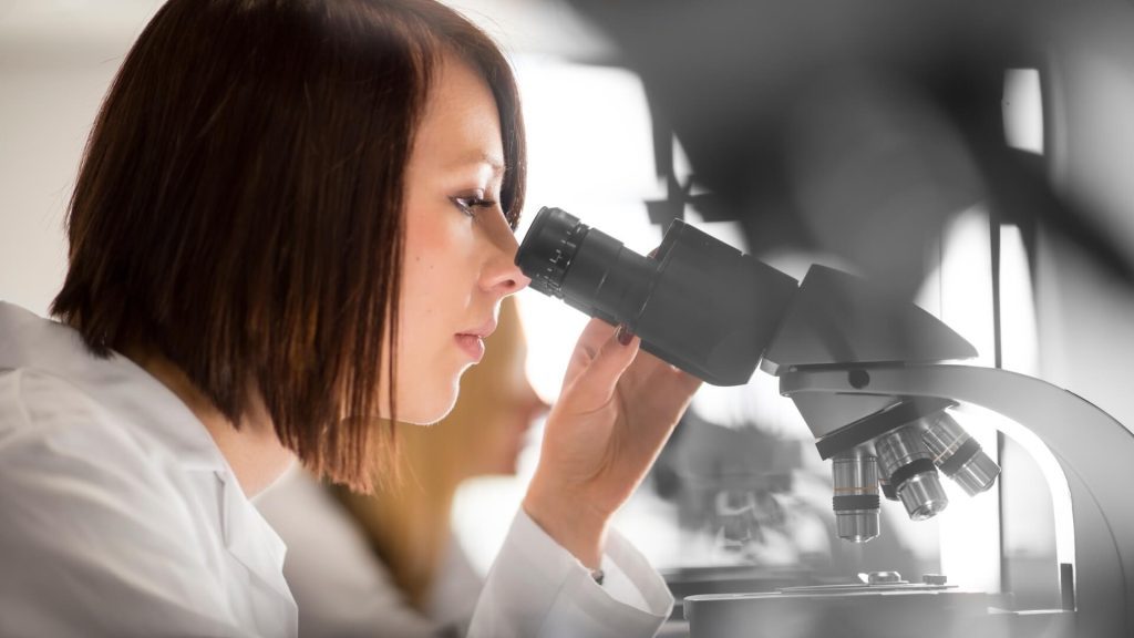 Student in white overalls in a laboratory looks into a microscope.