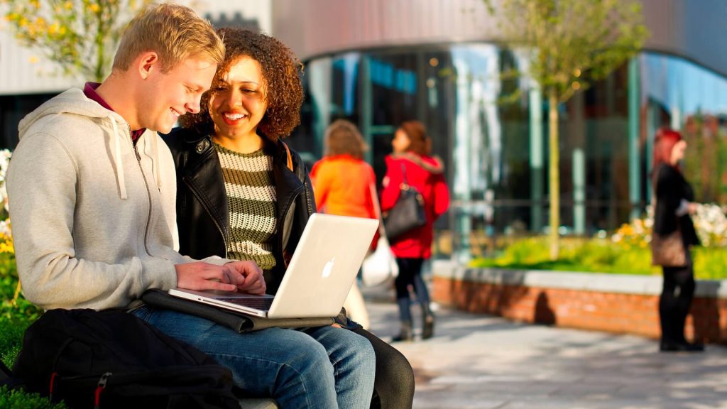 Two students sat talking outside the Hub, one using an Apple Mac device.
