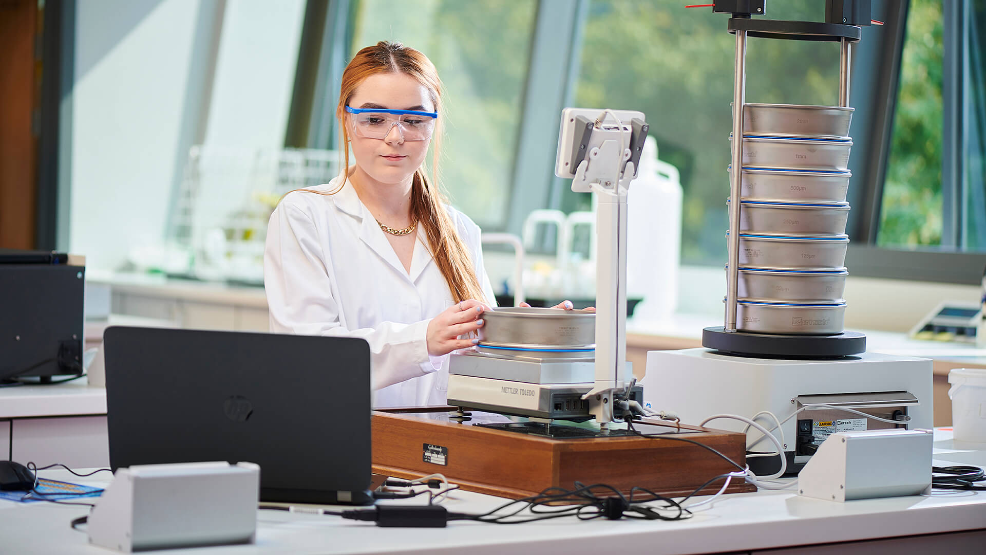 A student consults a laptop which is recording the mass of a sieve that is being weighed on a balance in preparation for particle size analysis using automated equipment and software.