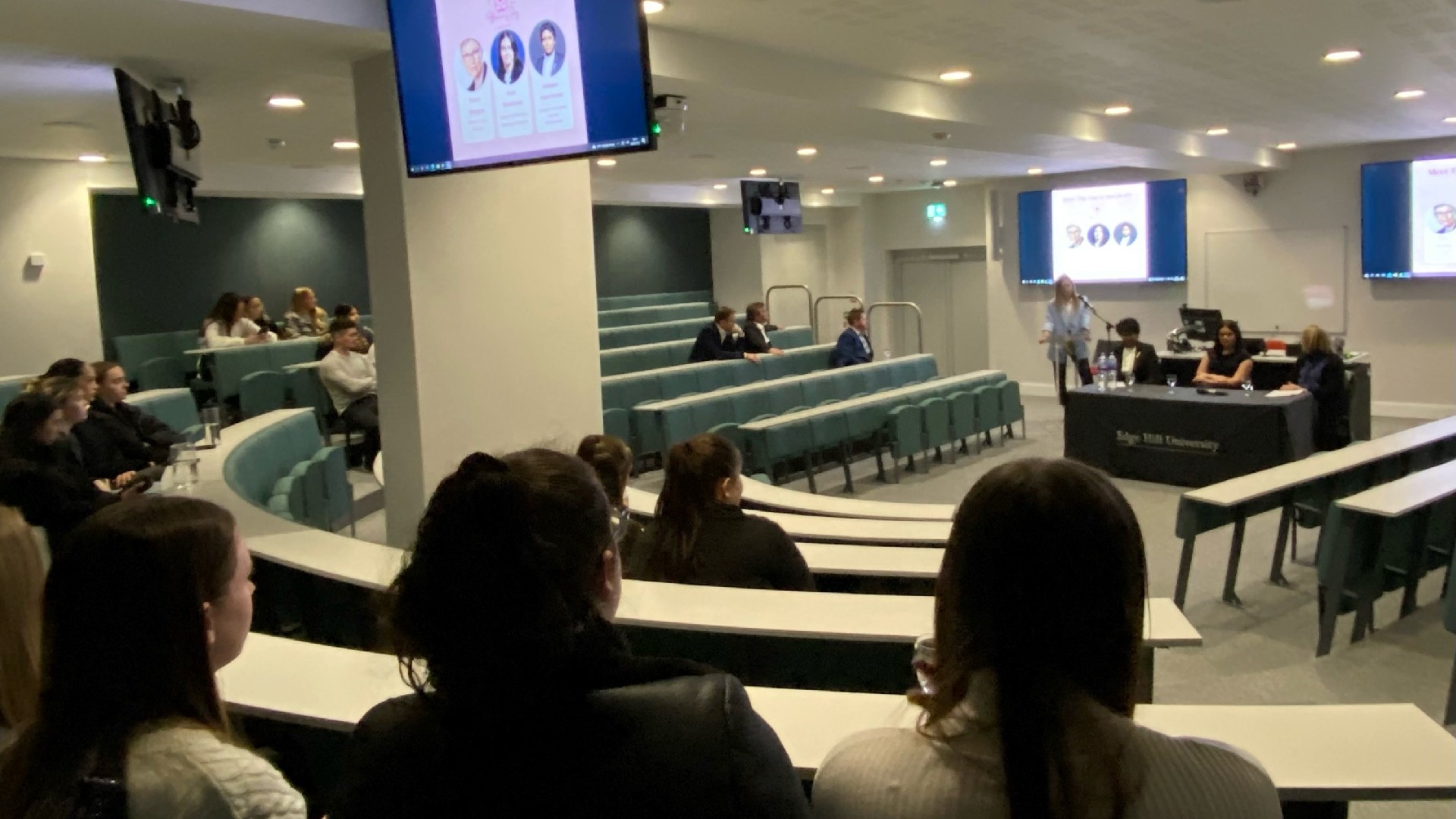 An image of the panel in session, multiple people are sat in a circle around a desk where one person is stood next to other people who are sat down.