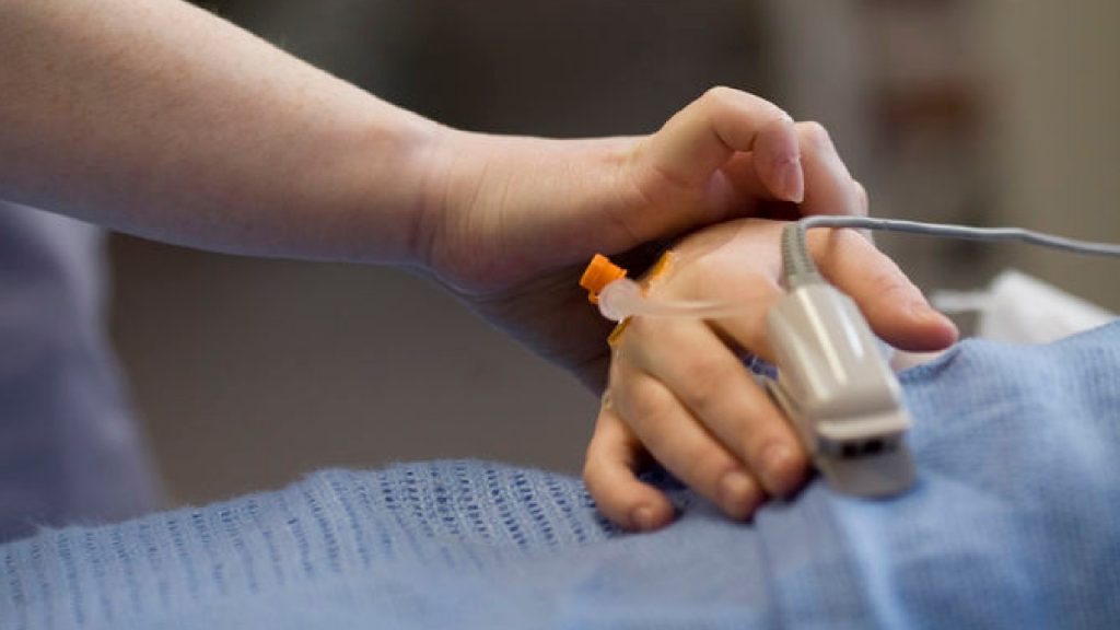 A nurse is holding the hand of a patient. There is a pulse oximeter on the finger on the finger of the patient.