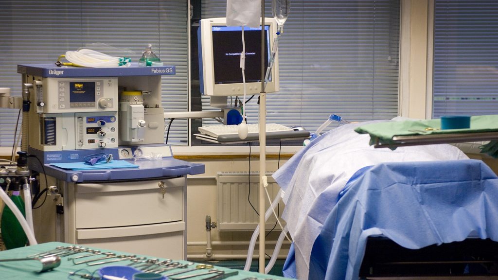 There is a variety of medical equipment in the photo. At the forefront of the image there are various medical tools on a green tray, behind these is a set of machinery and an operating table.