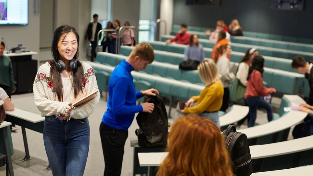 Two students talking to each other in a lecture theatre. There are other students in the background packing their things in the background, ready to leave the lecture theatre