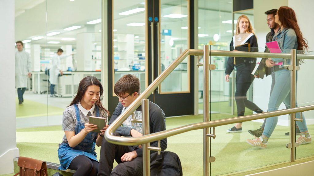 Two students are sitting on steps in the Biology building, looking at an iPad together. There are students walking past in the background. There is a lab with large glass windows in the background where students are working in lab coats.