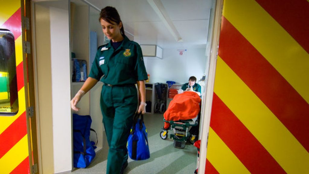 2 paramedics are inside an ambulance, one is walking out of the doors, the other is at the back of the ambulance, there is a patient on the bed within the ambulance.