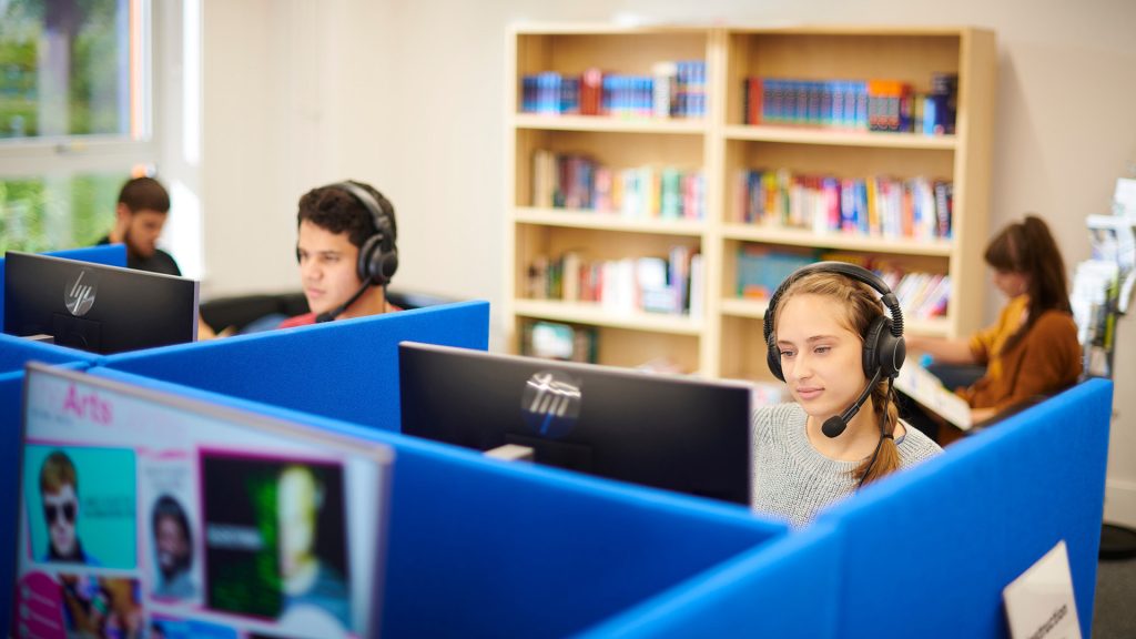 Two students sit in booths in front of computers with ear phones in