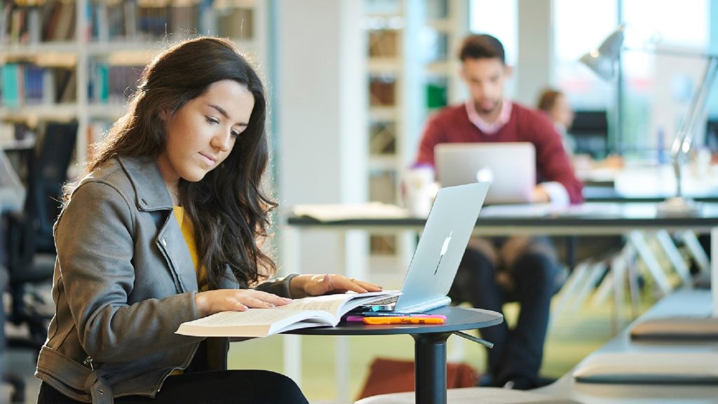 A student reads a book while working on her Apple Macbook at a table in the University library in the Catalyst building.
