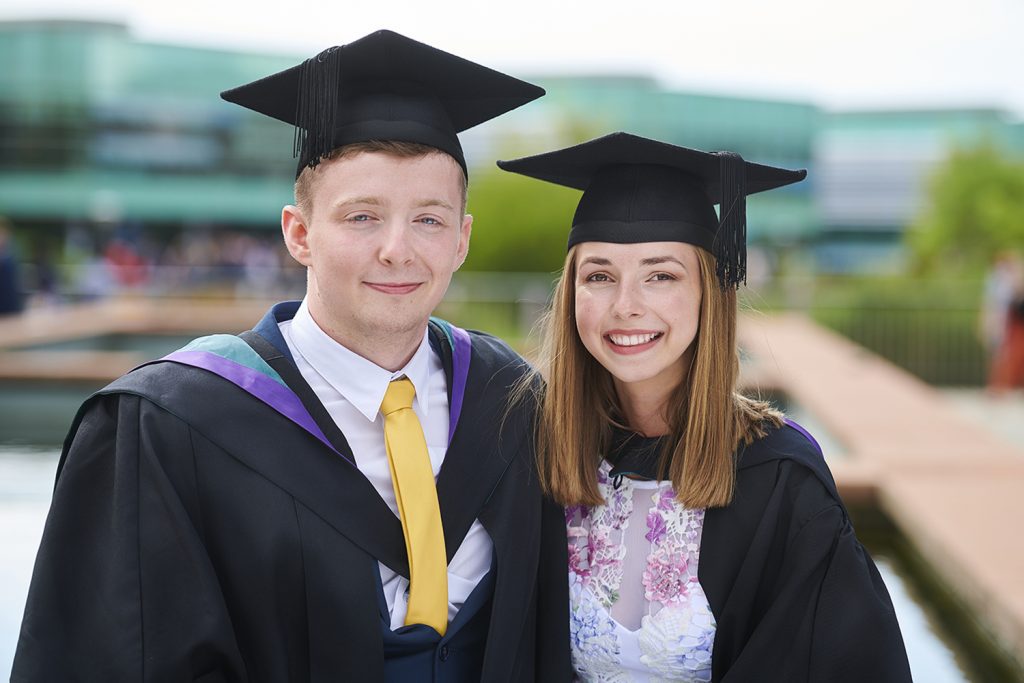 Two graduates, wearing their caps and gowns, stand on the piazza on the western side of campus.