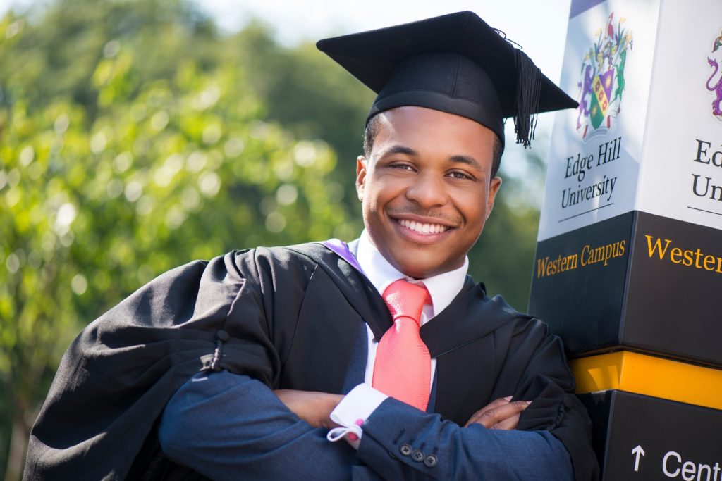 A graduate poses by a direction information sign on campus, wearing his graduation robe and mortar board.