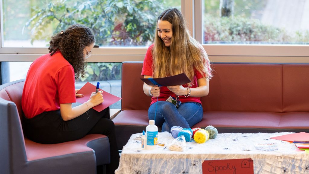 An image of two people sat on couches. They are both holding pieces of paper. On a table in front of them are various craft materials.