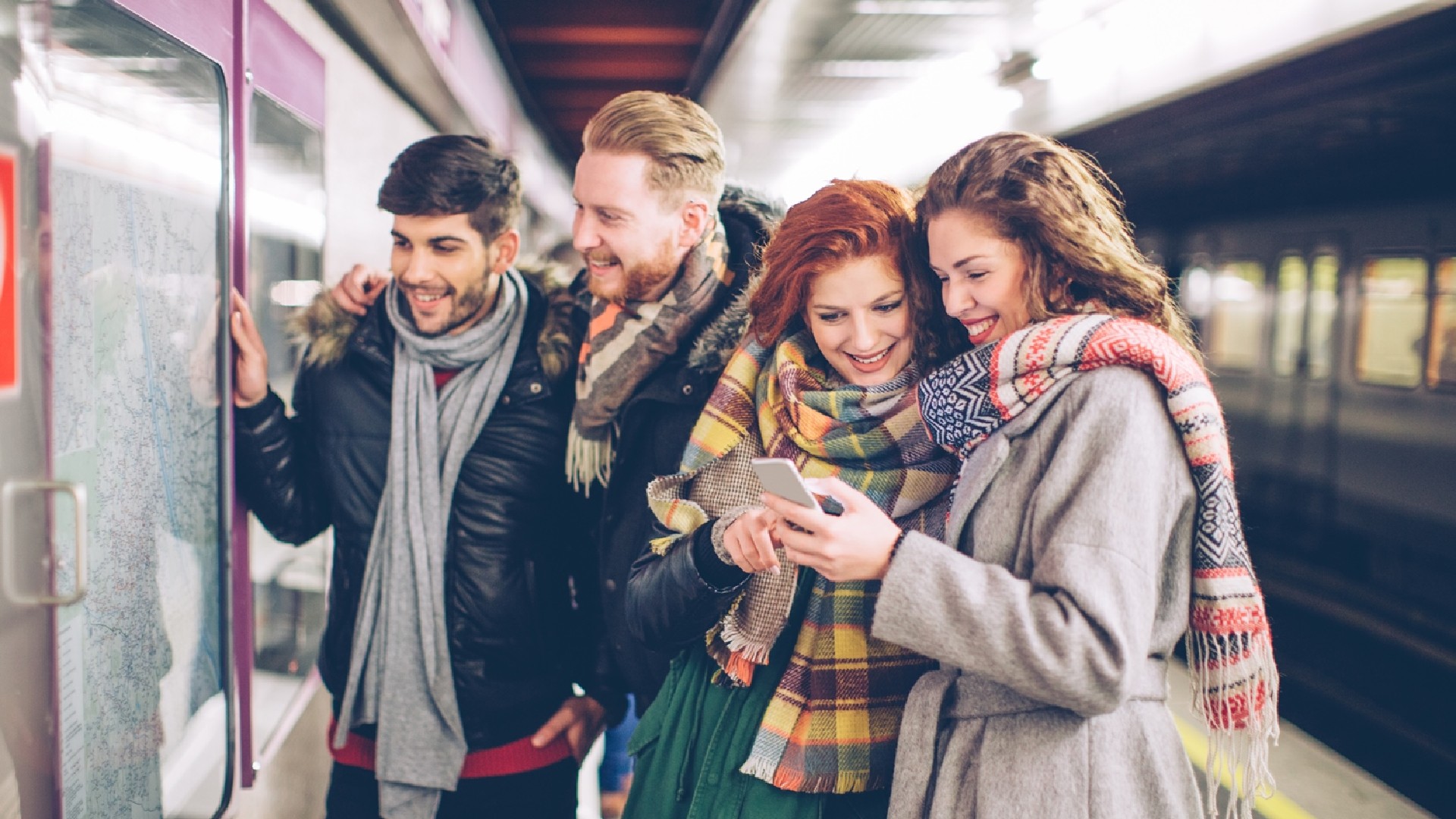 A group of people are in a train station. Two of the people are looking at a map on the wall, the other two people are looking at a mobile phone. In the background of the image there is a train waiting at the platform.
