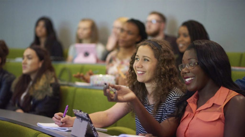 Two students are sat in a busy lecture hall smiling at the screen in front