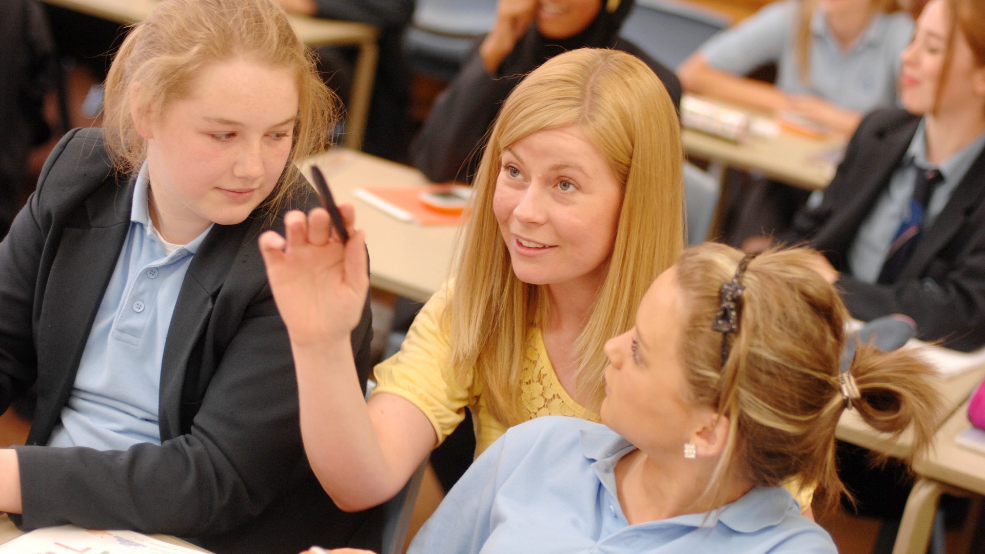 A teacher is sat in between two students pointing at something in front of that that is off-camera.