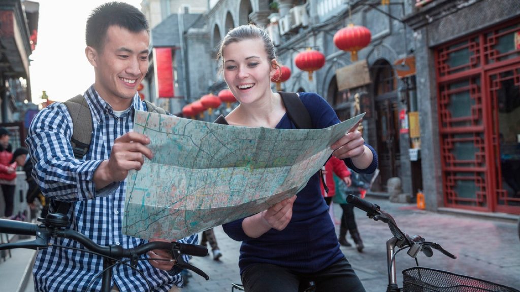 Two people are on bikes in the centre of a town. They are looking at a map that they are both holding.