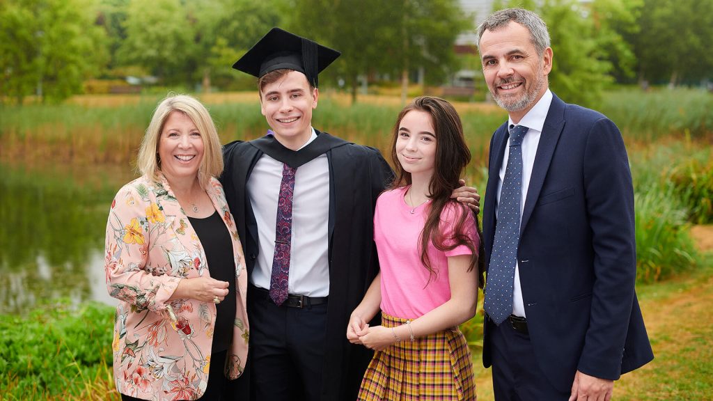 A graduate poses for a photo with his family next to the lake on the western side of campus while wearing his graduation robe and mortar board.