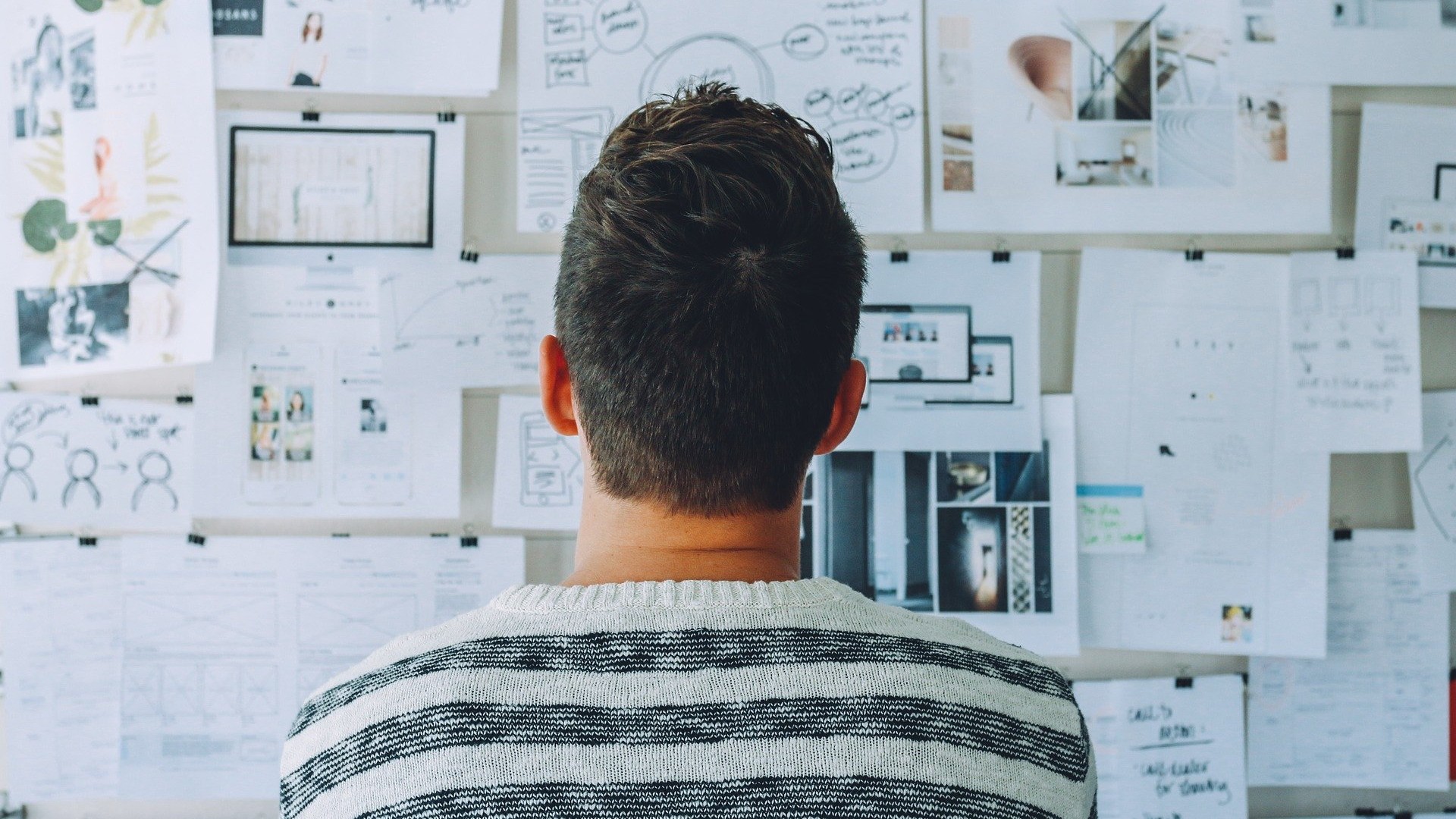 An image of a person stood looking at a wall that has multiple pieces of paper on and various drawings.