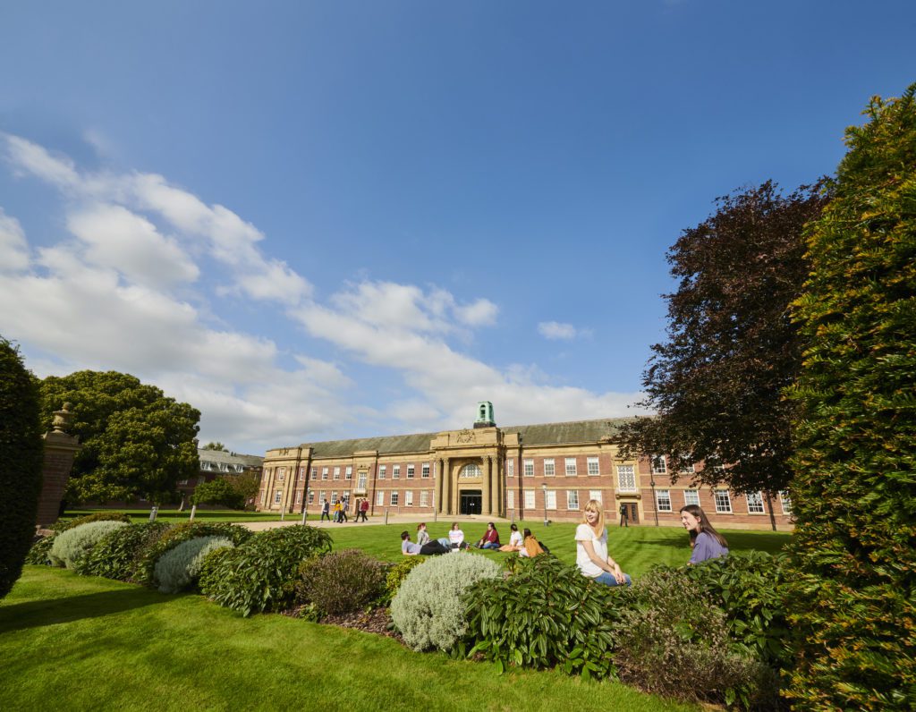 A picture of Edge Hill's Main Building. Students are sat on the grass outside surrounded by bushes and trees.