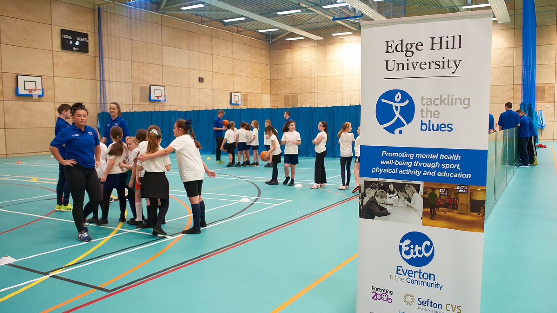 An image of a group of people stood in a group, next to them is a sign promoting the Edge Hill and Everton partnership.