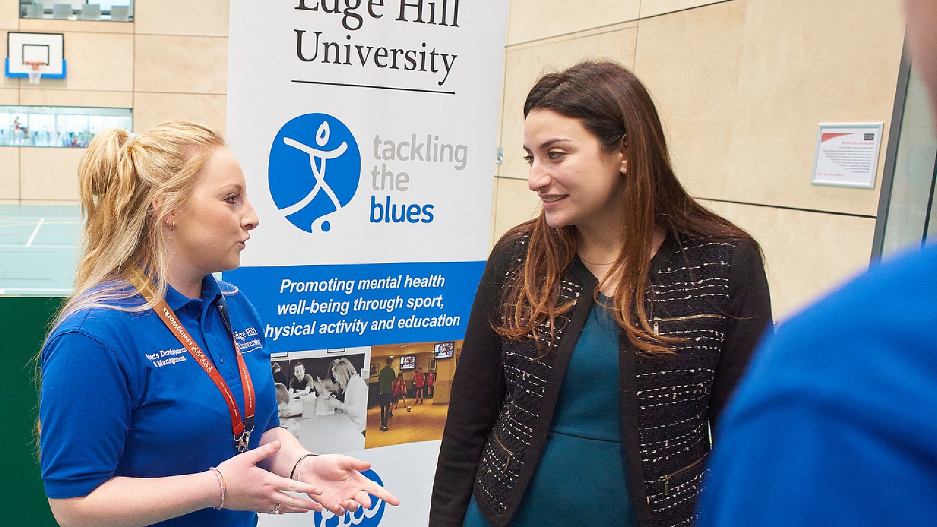 Two people are stood talking in front of a sign promoting the Edge Hill and Everton partnership. 