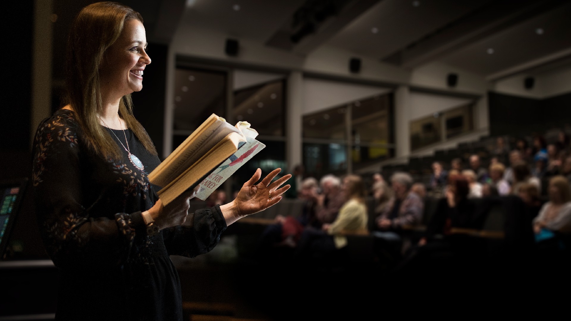 Cary Bray reading from her book on stage with a full audience in the row in front of her