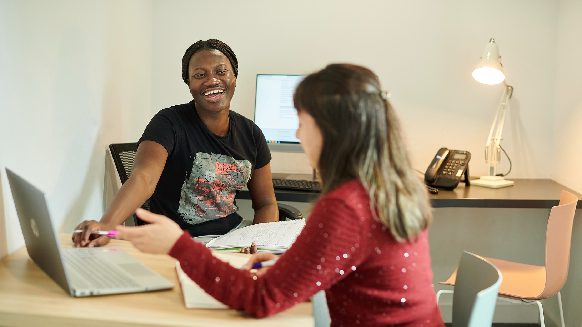 Two students laugh and talk in front of a laptop in a small room
