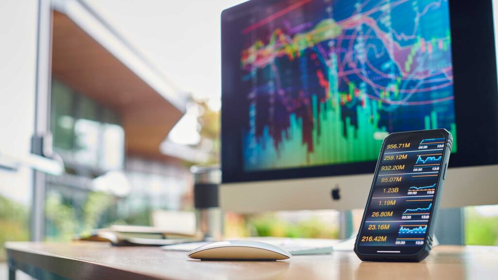 A smartphone on a desk showing market trends. An Apple iMac is in the background on the desk displaying data.