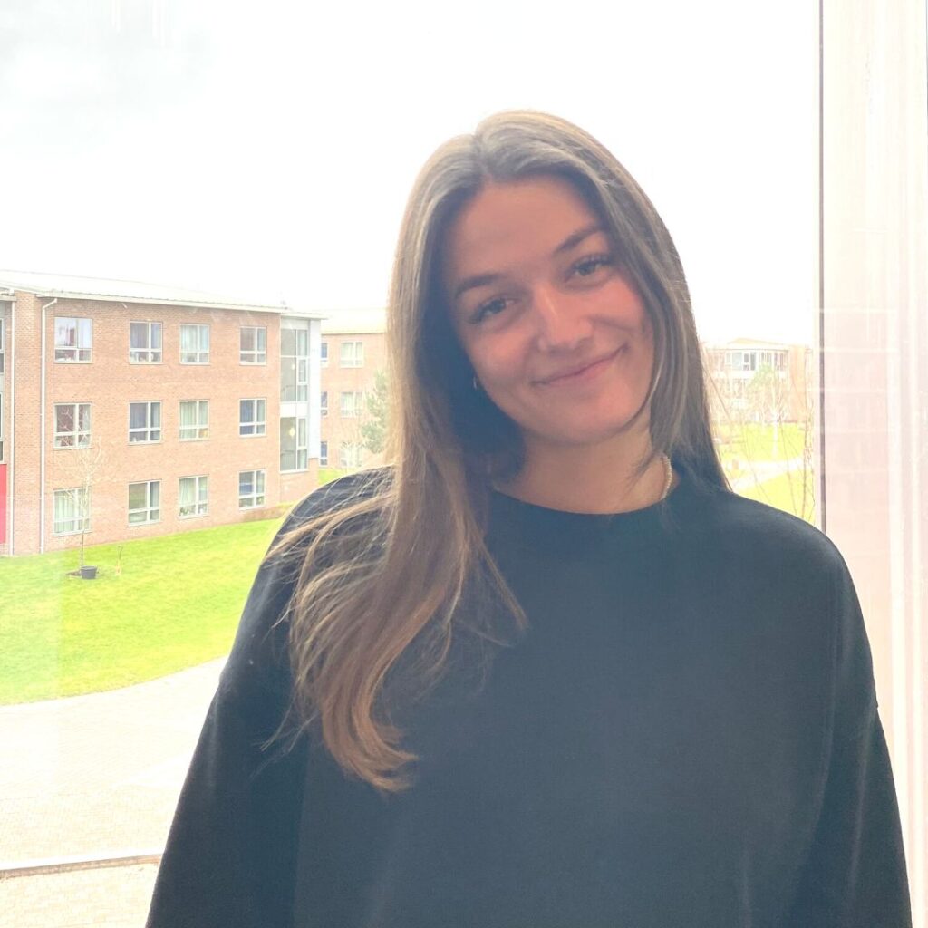 Law student facing the camera and smiling. stood at a window. university campus in the background