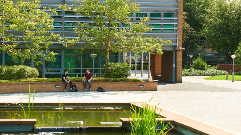 Image of a lake in front of the Geosciences building. Students sit on a nearby wall. / Geosciences