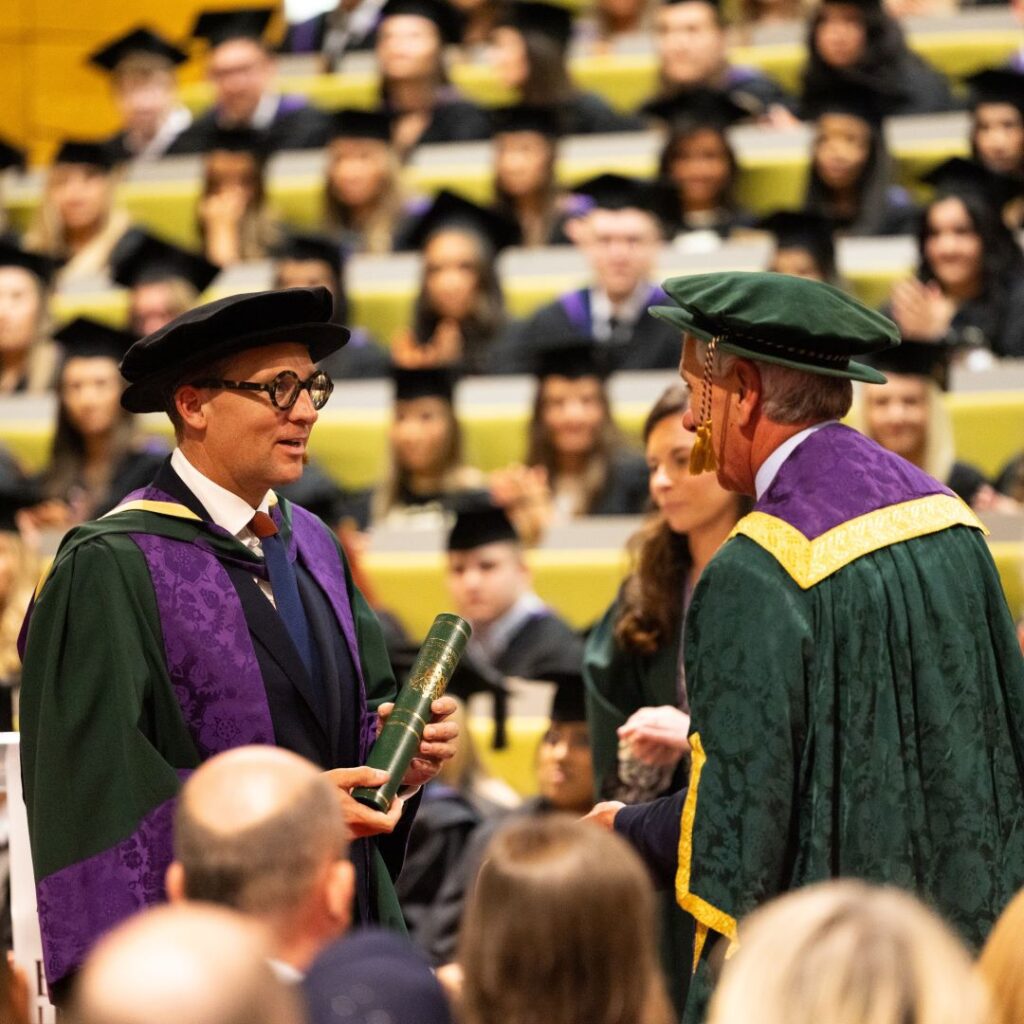 A picture of James Timpson collecting his honorary doctorate certificate from Vice-Chancellor John Cater. There is a large audience of students in the background.