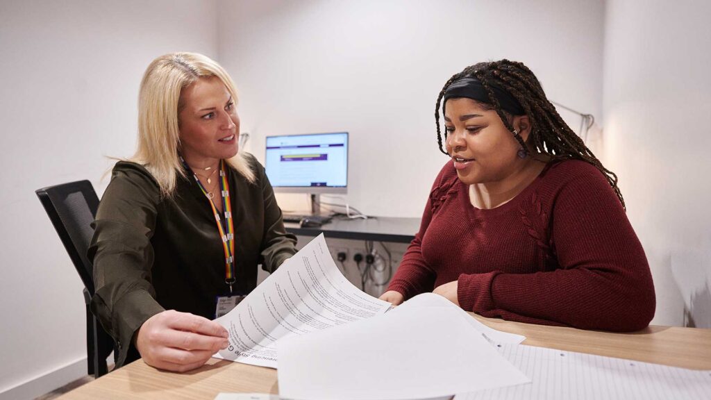 A student sat with a staff member in a Catalyst meeting room. They are reviewing paperwork together whilst talking.