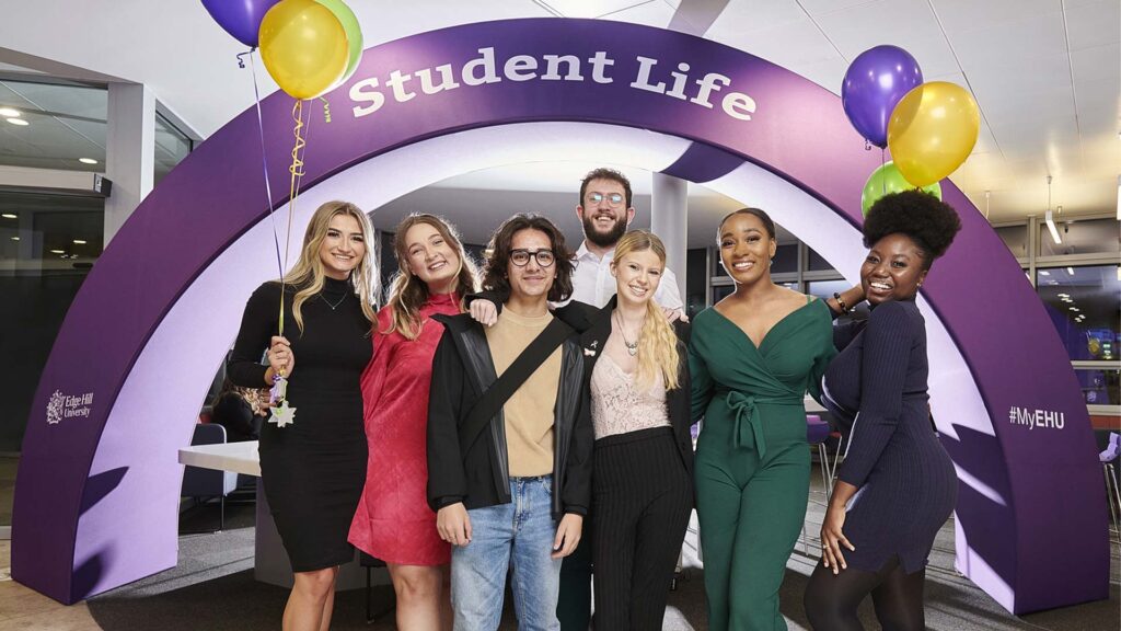 Image of students celebrating being awarded a scholarship. Seven students smiling stood beneath the 'Student Life' arch
