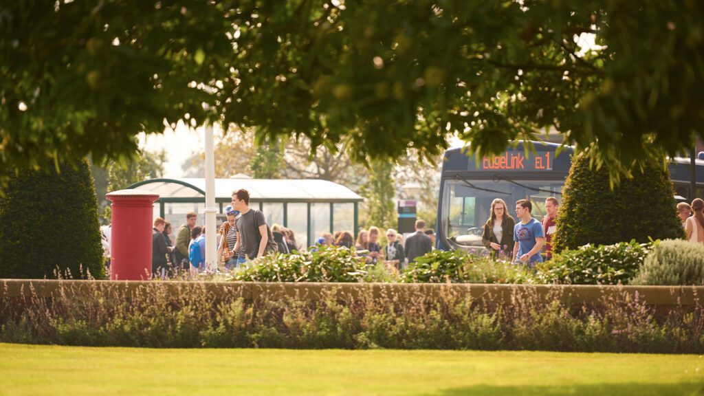 shot of the edgelink bus through the tree at the front of campus. lots of students walking round.