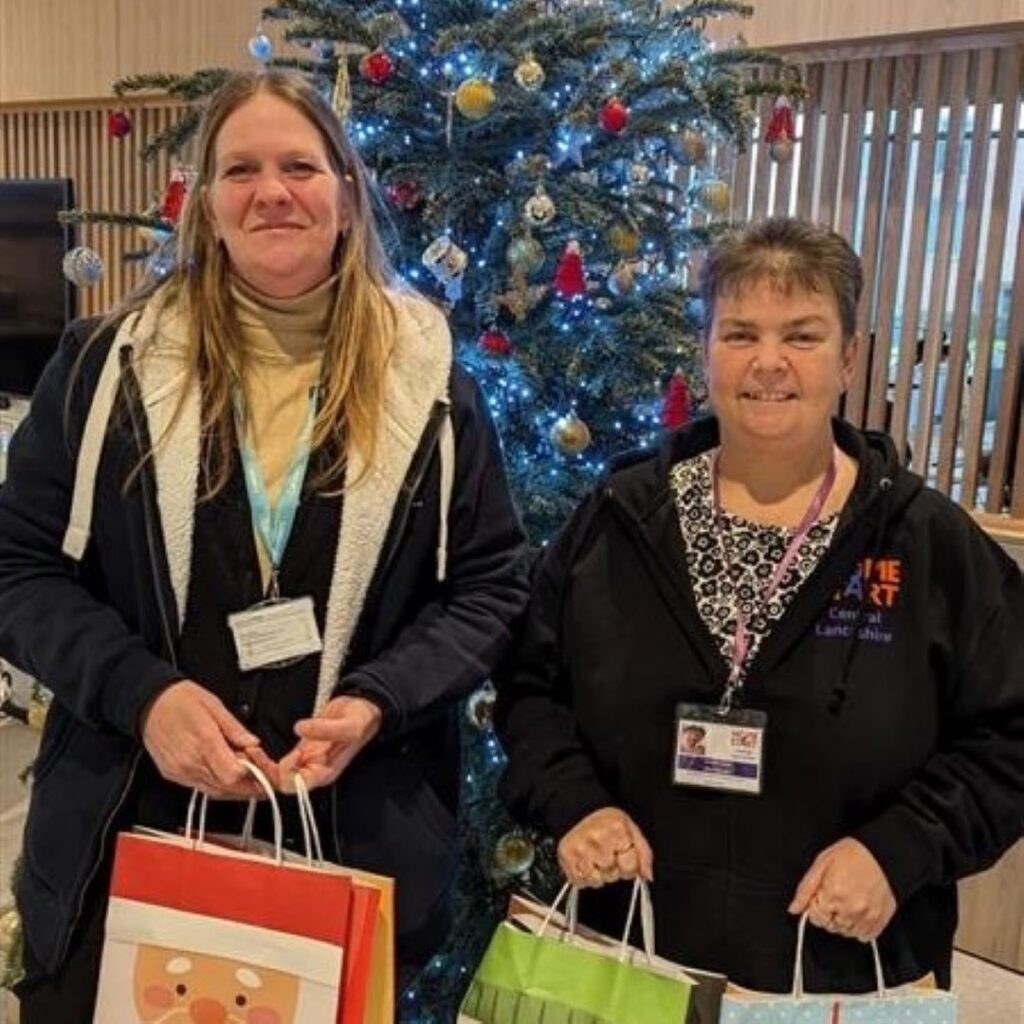 Two people stand in front of a Christmas tree with gift bags in their hands.