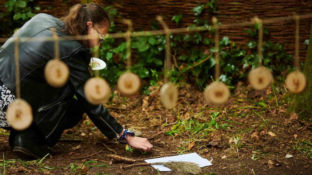 Forest School resources out of focus in foreground, a female teacher placing something on the ground in the forest.