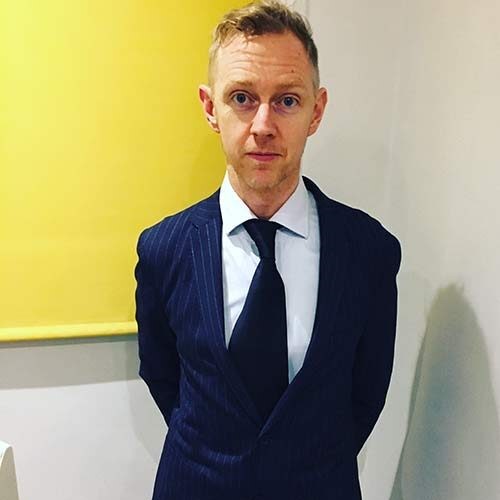 Tom Conaghan wears a suit and stands in front of a yellow blind.