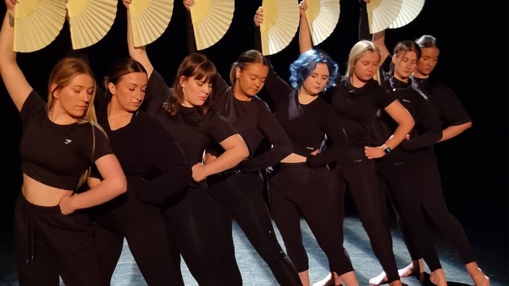 A group of dancers dressed in black outfits, holding yellow fans above their heads in a synchronized pose, performing on a dark stage with focused lighting.