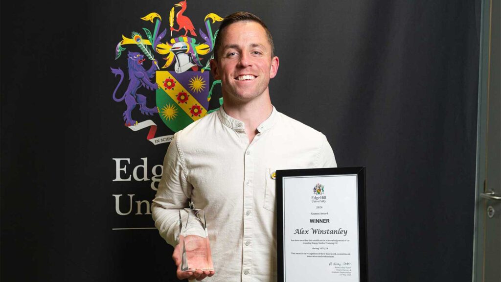 Alumni Alex Winstanley holding a glass award and certificate stood in front of a black backdrop showing the Edge Hill University crest