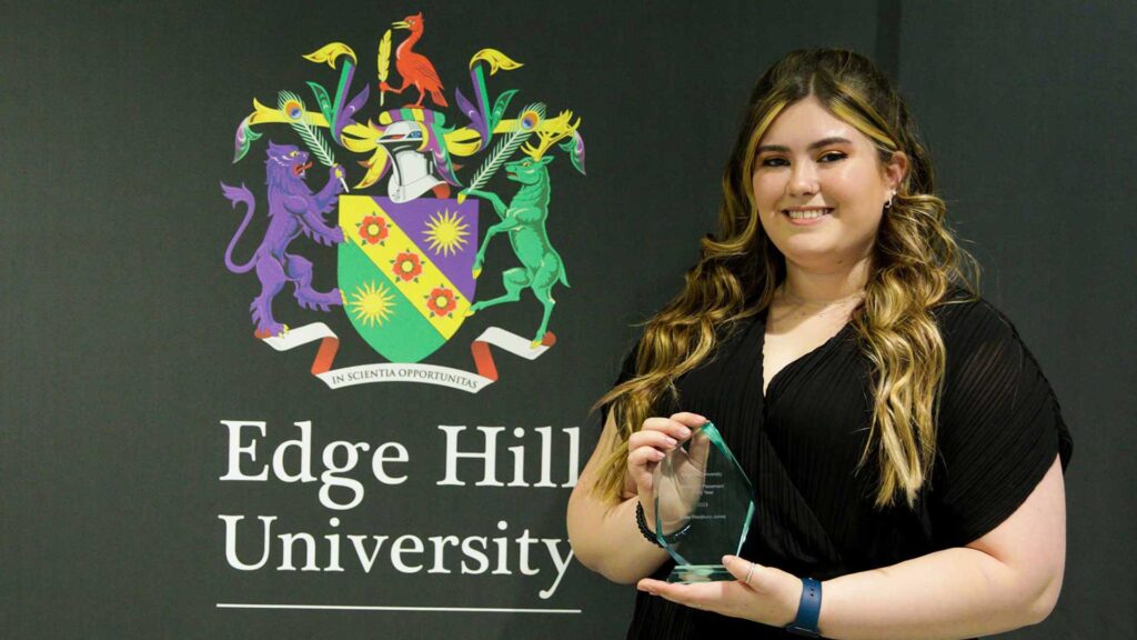 Student Elise Westbury-Jones holding a glass award stood in front of a black backdrop showing the Edge Hill University crest