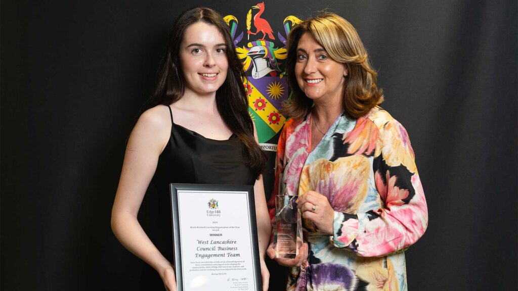 Two members of West Lancashire Council: Business Engagement Team holding a glass award and certificate stood in front of a black backdrop showing the Edge Hill University crest