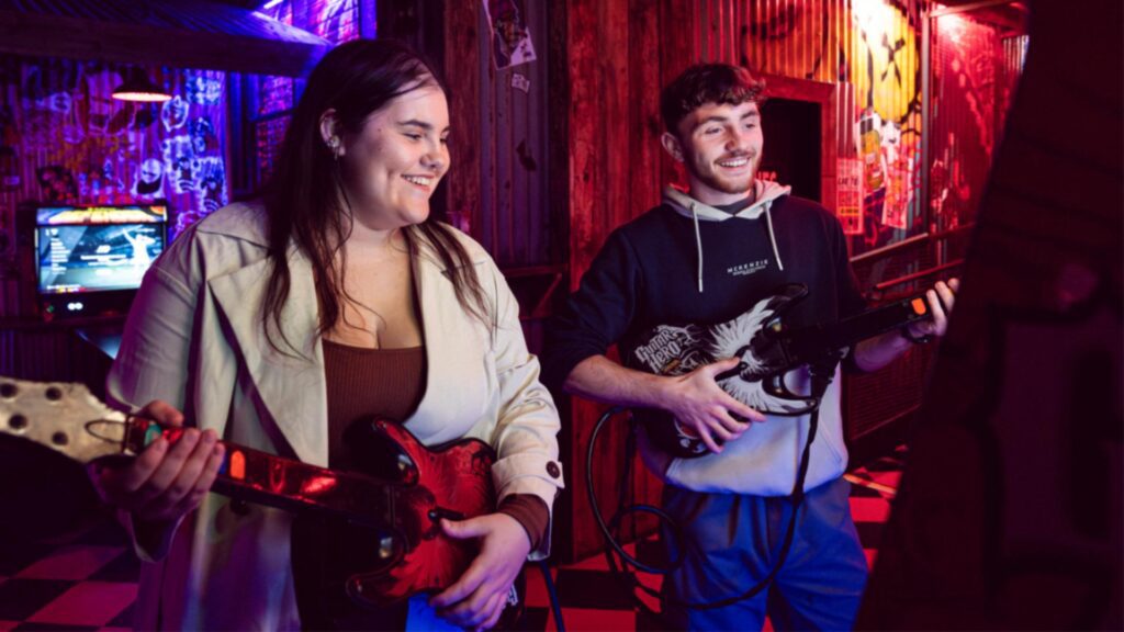 Two students play Guitar hero together in a retro gaming bar.