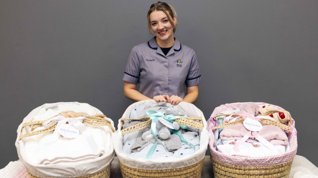 Athalia wears her student midwife uniform and stands behind a trio of moses baskets filled with baby clothes and nappies.