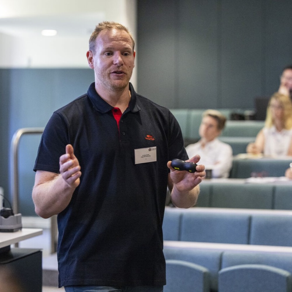Dr Sven Batke wearing a blue sweatshirt, standing up and addressing an audience at an event.