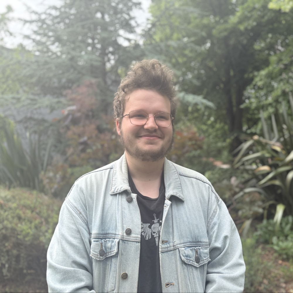 student Alex, standing outside with greenery behind him, wearing black t-shirt and denim jacket.